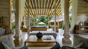 Un espacio interior amplio con pilares de madera, una zona de estar central con una mesa, y una piscina visible al fondo.