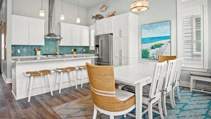 Une cuisine moderne avec des meubles blancs, un îlot central doté de tabourets, des appareils en acier inoxydable et une grande fenêtre donnant sur la plage.