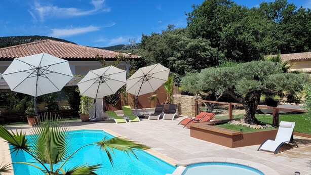 A swimming pool with lounge chairs and umbrellas, surrounded by a fenced area with trees and a building in the background. - --- GENEVRIER cottage --- (Quissac)
