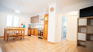 A kitchen with wooden cabinets, a dining table with chairs, a clock on the wall, and a TV on the wall. - --- GENEVRIER cottage --- (Quissac)