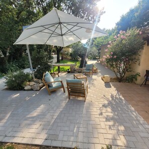 A patio with a table and chairs under a large umbrella, surrounded by greenery and a stone wall. - --- GENEVRIER cottage --- (Quissac)