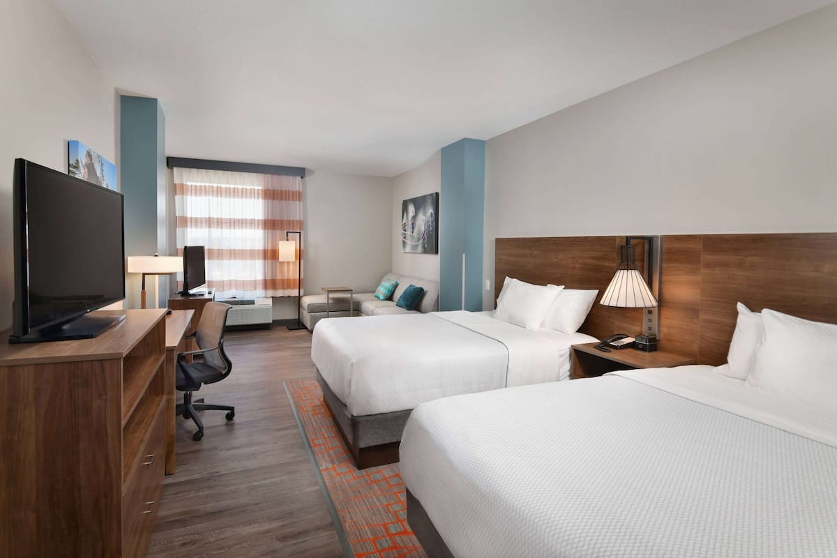 A hotel room with two beds, a desk with a TV, a chair, a lamp, and a window with curtains.