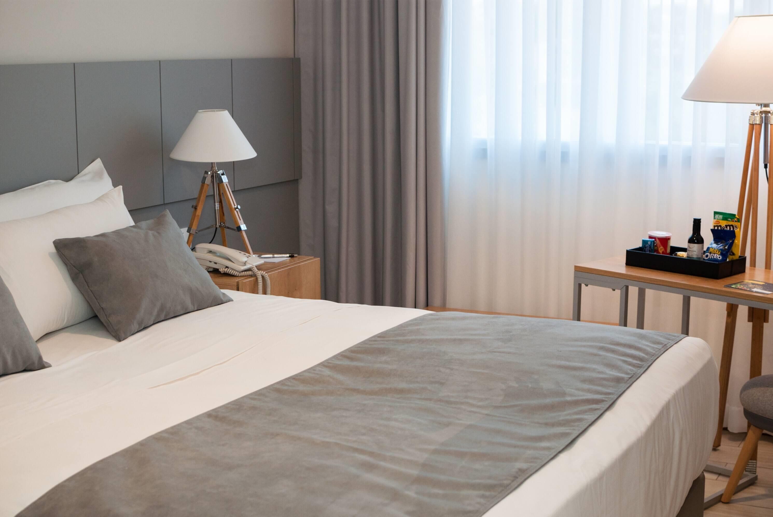 Une chambre d’hôtel avec un grand lit, une table de chevet avec une lampe, un téléphone et une petite table avec des en-cas et une bouteille.