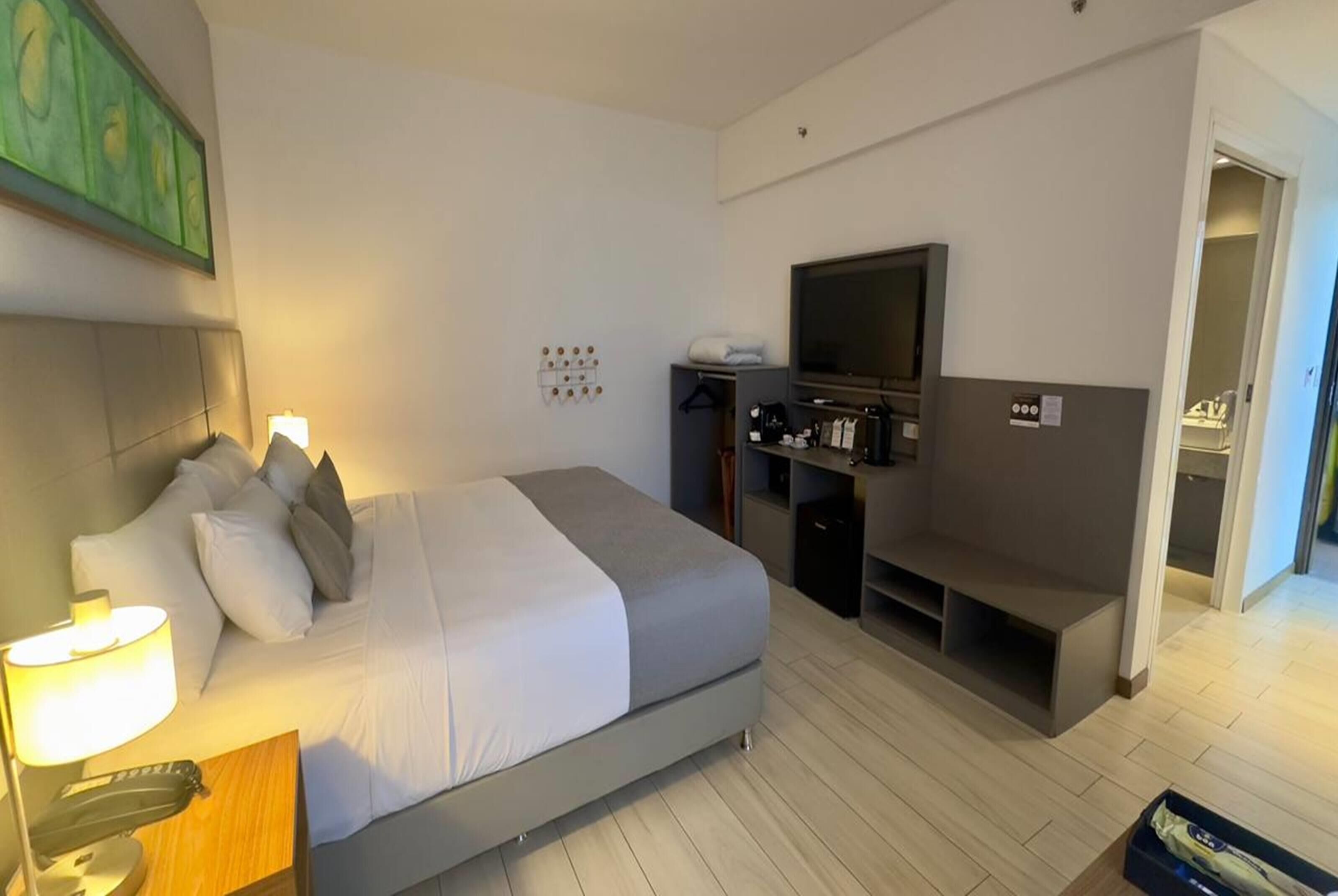 Quarto de hotel moderno com cama, luminária de cabeceira, televisão e um pequeno espaço de estar.