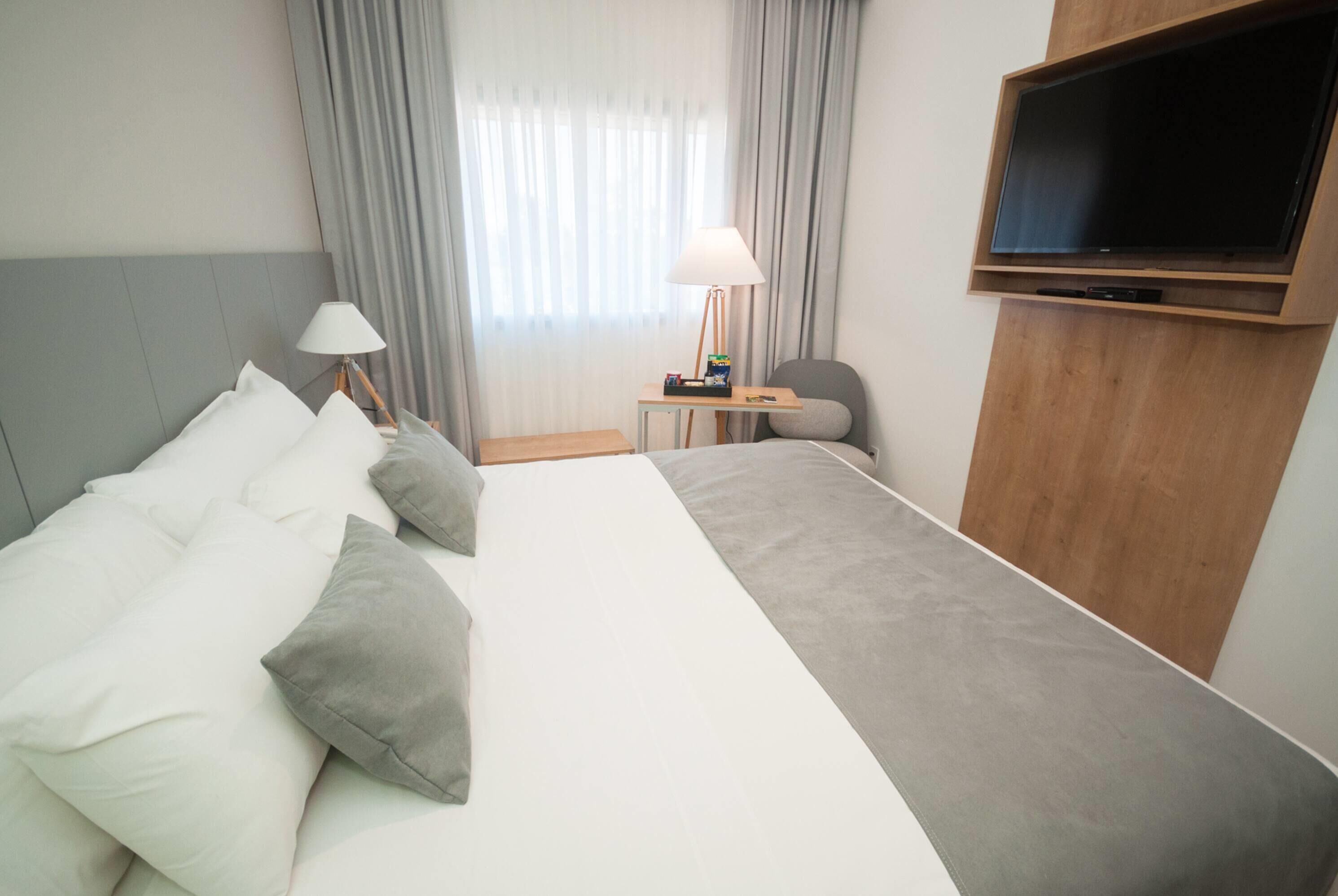 Ett hotellrum med en stor säng, en tv som är monterad på väggen, ett skrivbord med en lampa och en stol.