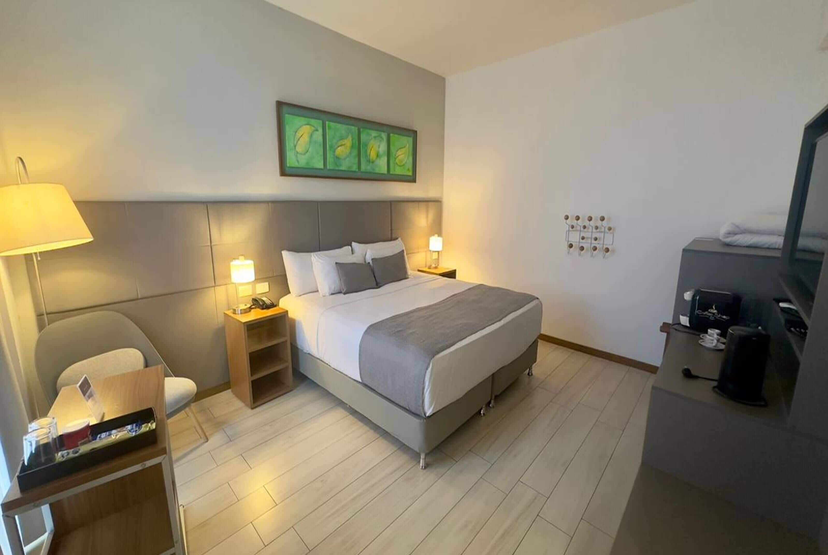 A hotel room with a bed, bedside tables, a desk, a chair, a TV, and a coffee machine.