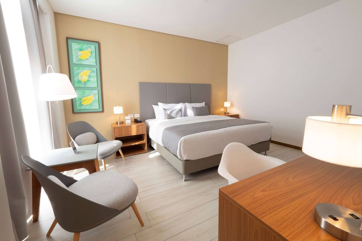 Ein Hotelzimmer mit einem Bett, zwei Nachttischen mit Lampen, einem Schreibtisch mit Stuhl und einem Kunstwerk an der Wand.