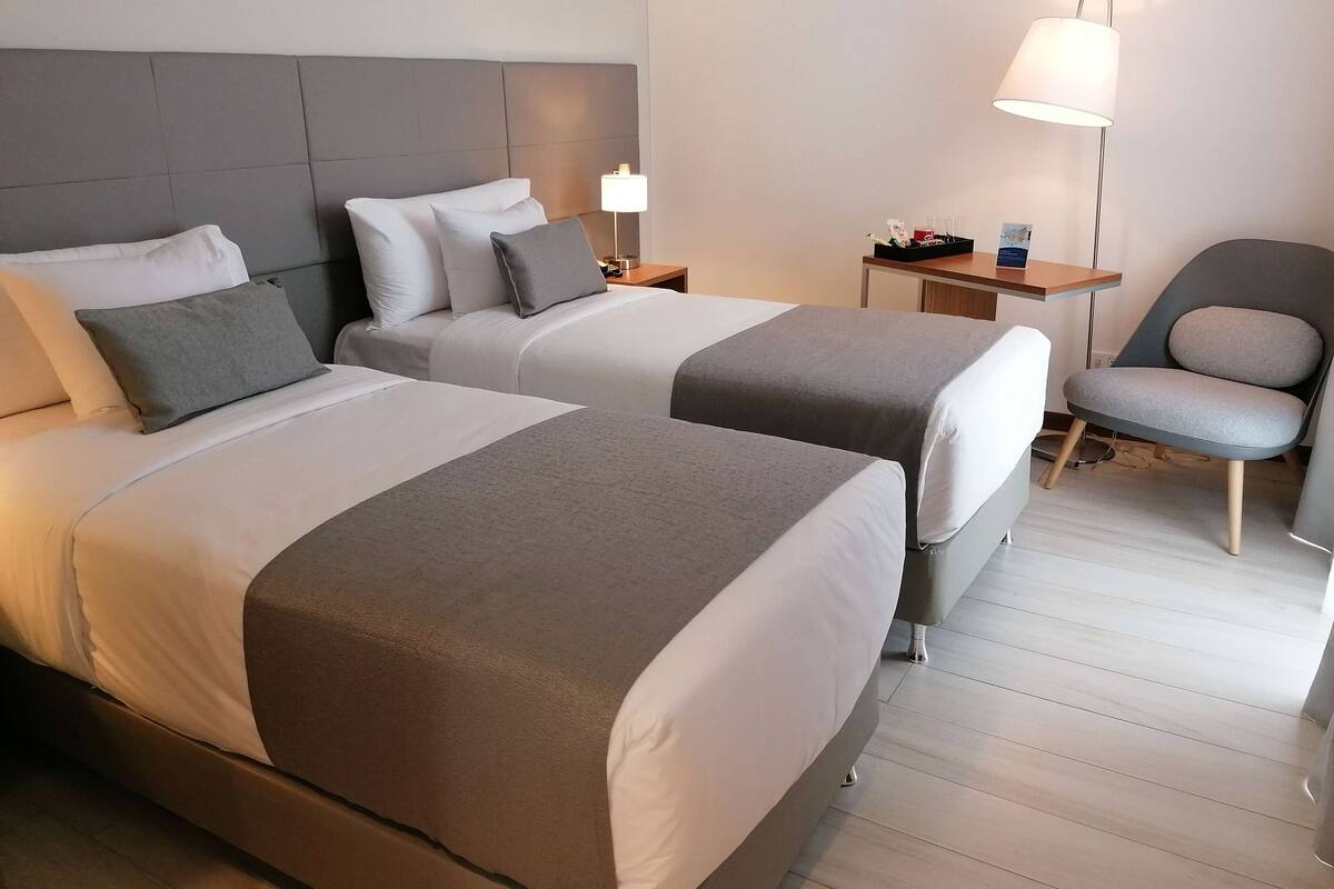Ein Hotelzimmer mit zwei Betten, einem Sessel, einem Nachttisch mit Lampe und einem kleinen Tisch mit Karte und Vase.