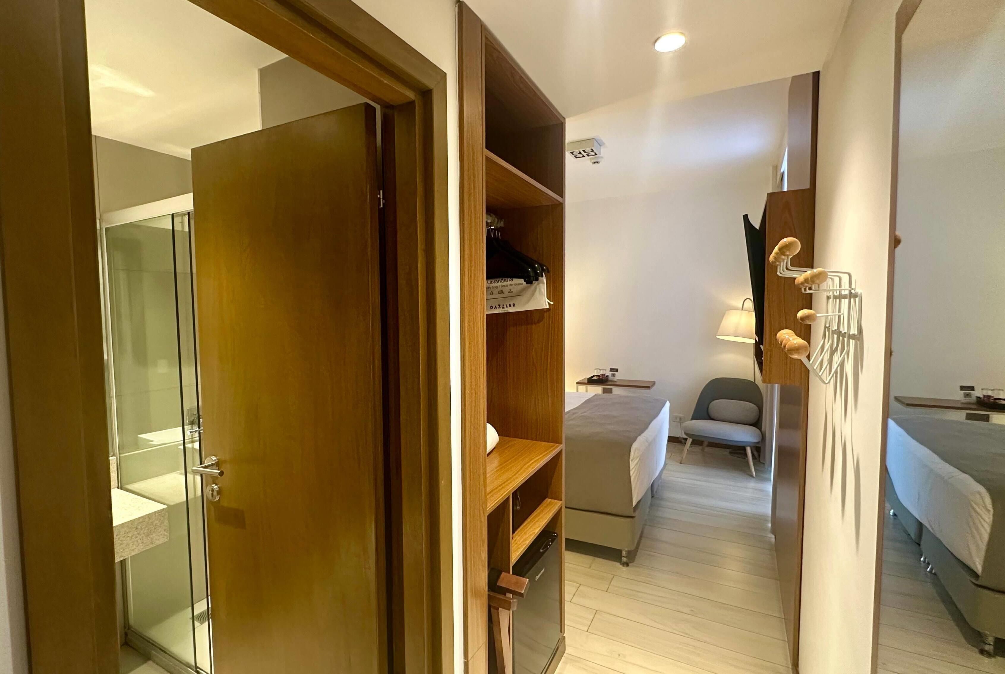 A hotel room with a wooden wardrobe, a bed, a bedside table, a lamp, and a shower area.