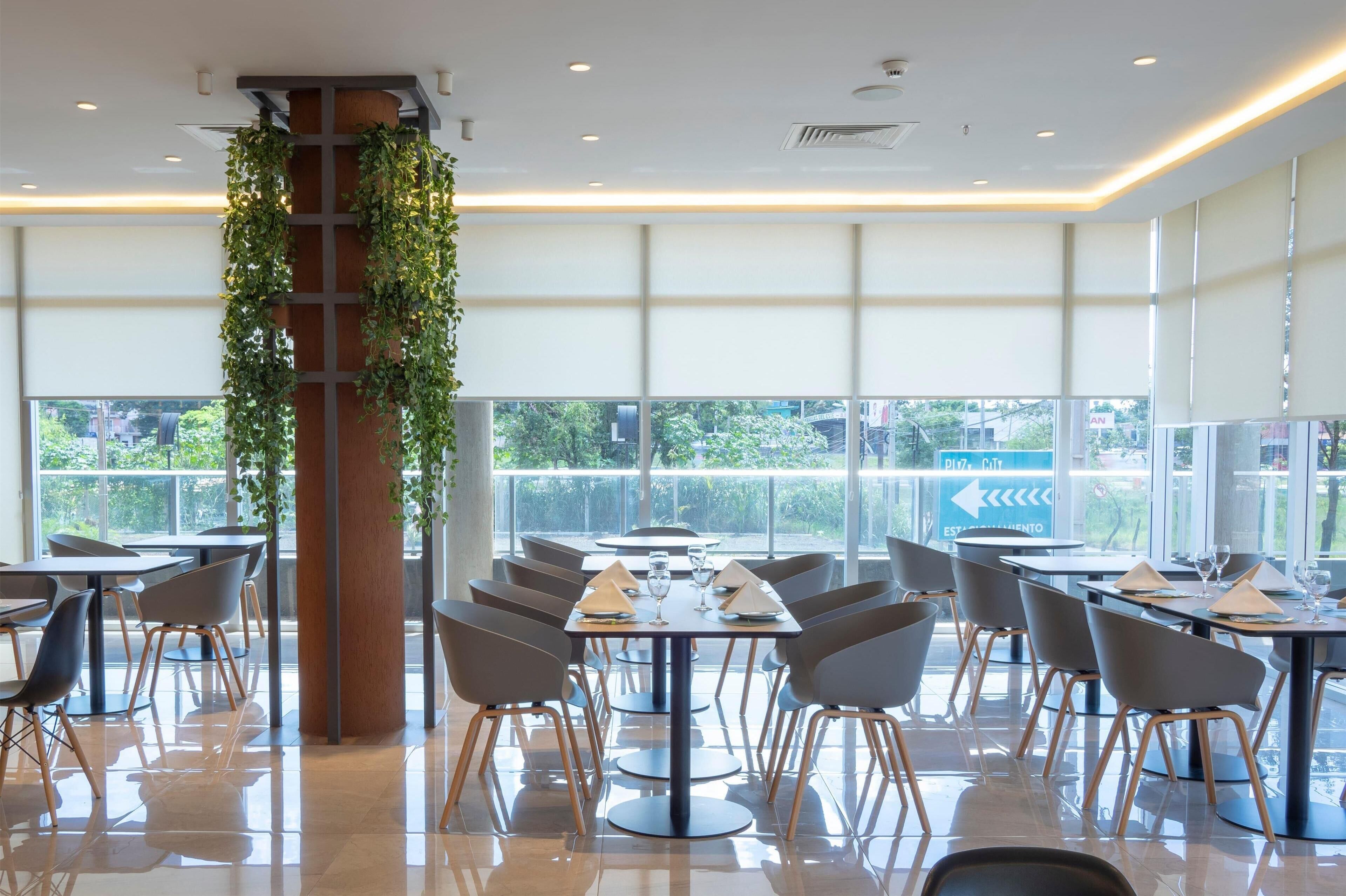 A modern restaurant with a high ceiling, large windows, and a central column adorned with greenery. Tables are neatly arranged with chairs, and the floor is polished.