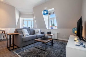 Una sala de estar moderna con un sofá gris, una mesa de centro y un televisor. Hay dos ventanas con cortinas, una lámpara de techo y una mesita de noche con una lámpara.