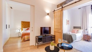 Une chambre d’hôtel moderne avec un grand lit, une télévision, une petite table avec une plante et une porte coulissante donnant sur une autre pièce.