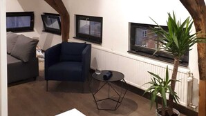 A loft-style room with exposed wooden beams, a grey sofa, a blue armchair, a small round table, and a potted plant. - Flat at Grand Place. (Brussel)