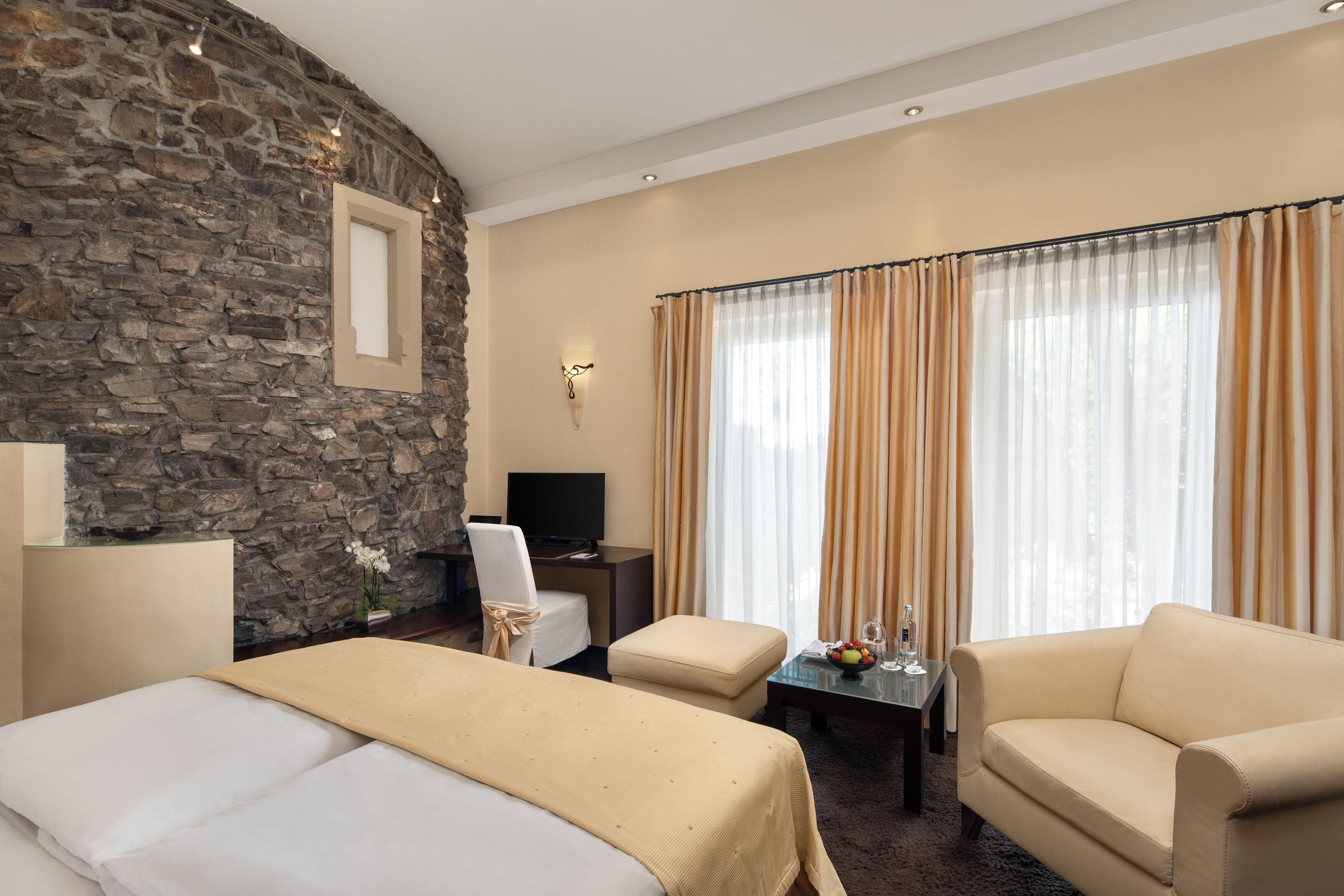 A hotel room with a stone wall, a bed, a sofa, a TV, and a window with curtains.