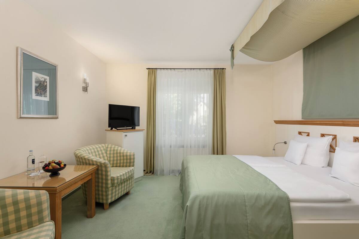 Habitación de hotel con cama, una silla, una mesita, televisor y ventana con cortinas.