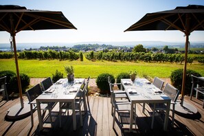 An outdoor dining area with tables and chairs, surrounded by a vineyard and a view of a town in the distance.
