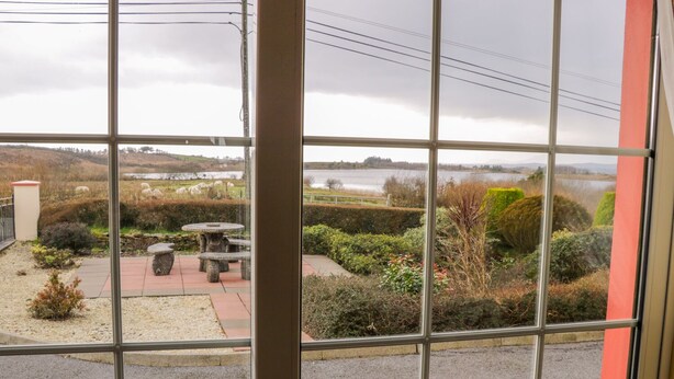 A view through a window of a garden area with a stone table and benches, a pathway, and a lake in the background. - Ellen's Cottage (Mountcharles)