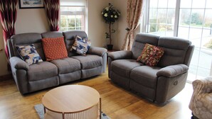 A living room with a grey sofa, a grey armchair, and a round wooden coffee table. There are two windows with curtains, a potted plant, and a framed picture on the wall. - Ellen's Cottage (Mountcharles)
