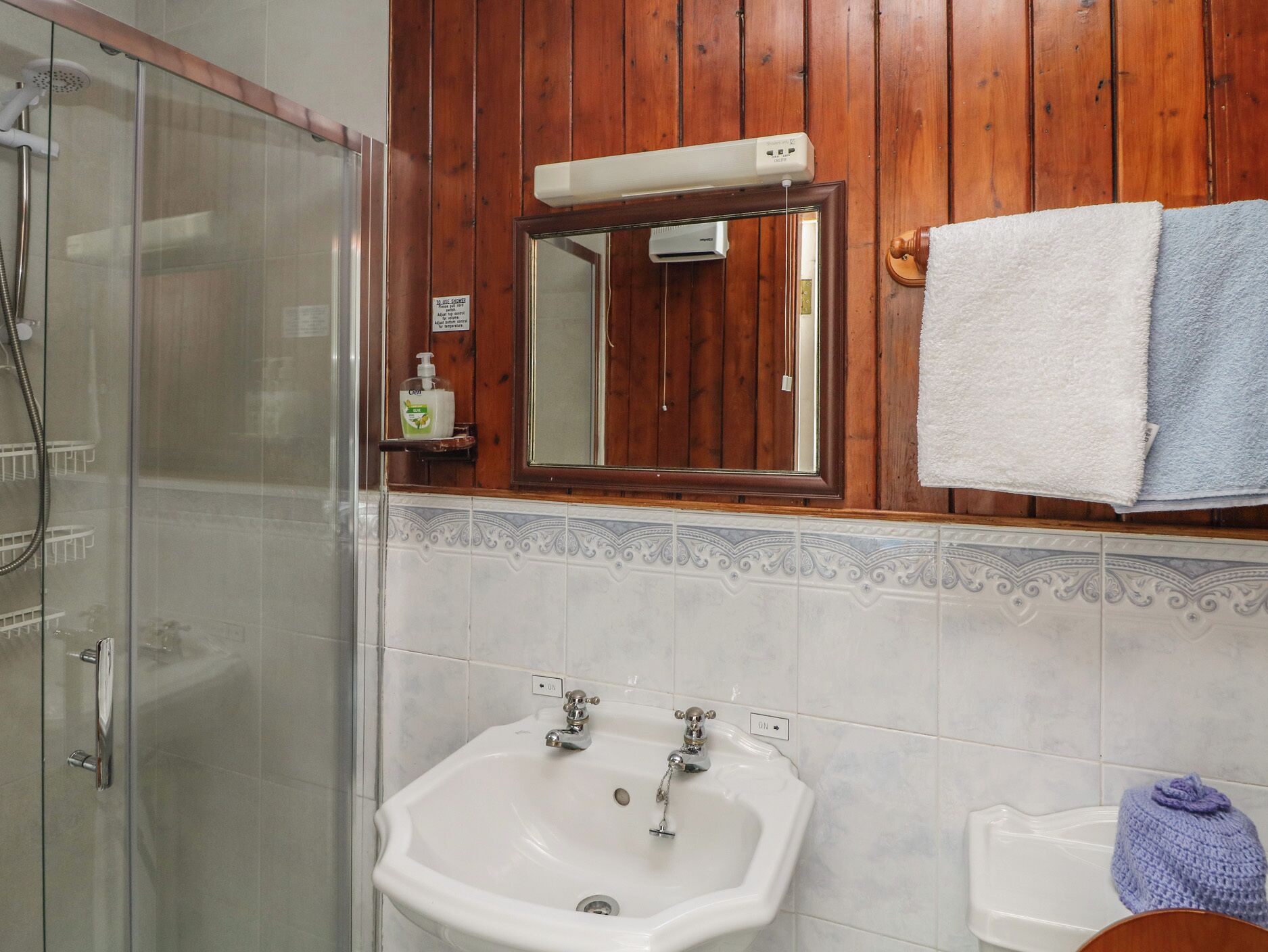 Un baño con cabina de ducha de vidrio, un lavamanos con dos grifos, un espejo sobre el lavamanos y un perchero con una toalla blanca y azul.