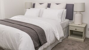 A neatly made bed with white bedding and a gray throw blanket. There is a bedside table with a lamp and a drawer. - Witts End (Redcar)