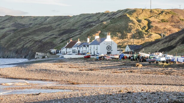 A coastal building with a prominent wheel on the facade, situated on a pebbled beach with boats and a hilly backdrop. - Witts End (Redcar)