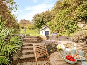 Un jardin avec un escalier en pierre menant à une petite maison, un banc en bois et une table sur laquelle se trouvent un saladier de fraises et un vase de fleurs.