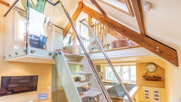 A modern interior with a glass staircase, wooden beams, a flat-screen TV, and a kitchen area. - Swallows Loft (Kingsbridge)
