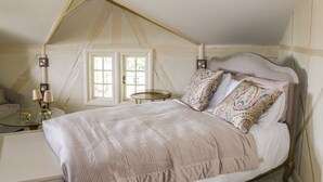 Ein Schlafzimmer mit schrägen Wänden, einem Bett mit gemusterten Kissen, einem Nachttisch mit Lampen und einem Fenster mit Vorhängen.