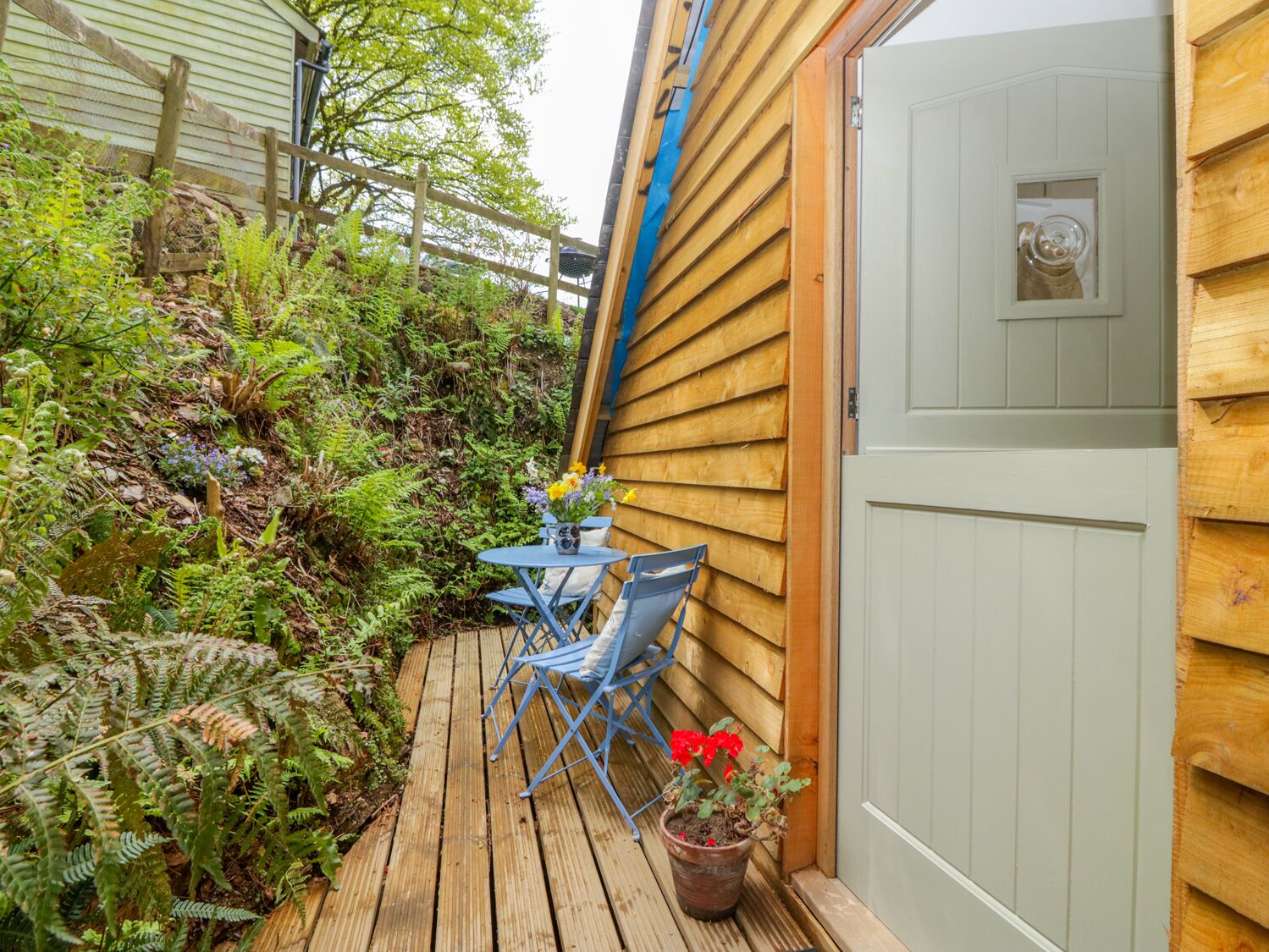 A wooden deck with a table and chairs, surrounded by lush greenery and ferns.