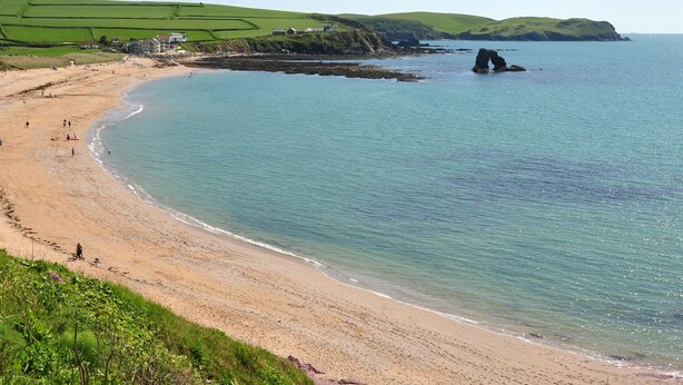 Una playa costera con vista clara al mar, una orilla arenosa y campos verdes al fondo.