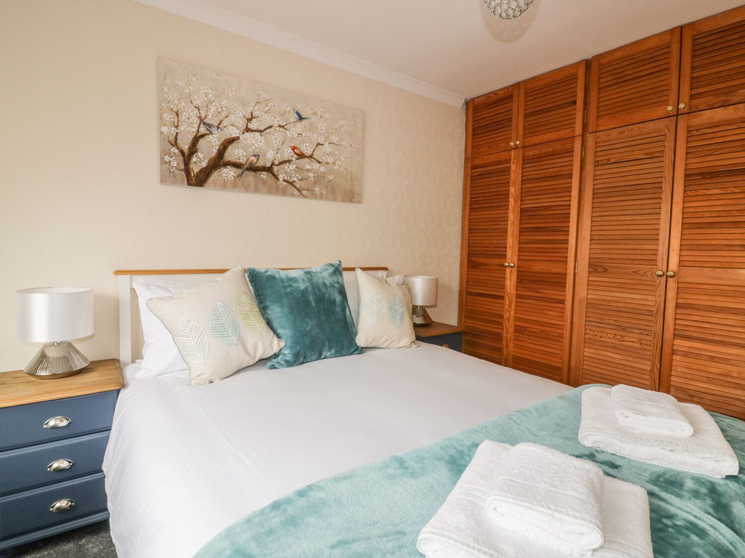 Quarto com cama, almofadas, mesa de cabeceira, abajur, guarda-roupa de madeira e pintura na parede.