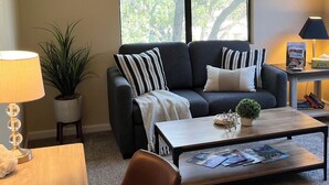 Een woonkamer met een bank, salontafel en een lamp. Een plant en een raam met zonwering.