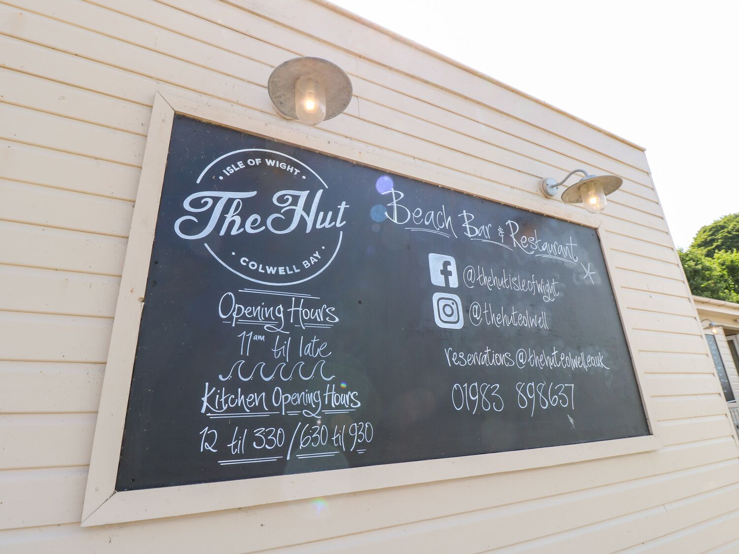 A chalkboard sign for The Hut, a beach bar and restaurant, with opening hours and contact information.