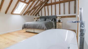 A bedroom with a large white bathtub, a bed with grey bedding, wooden beams on the ceiling, and a small bedside table with a lamp.
