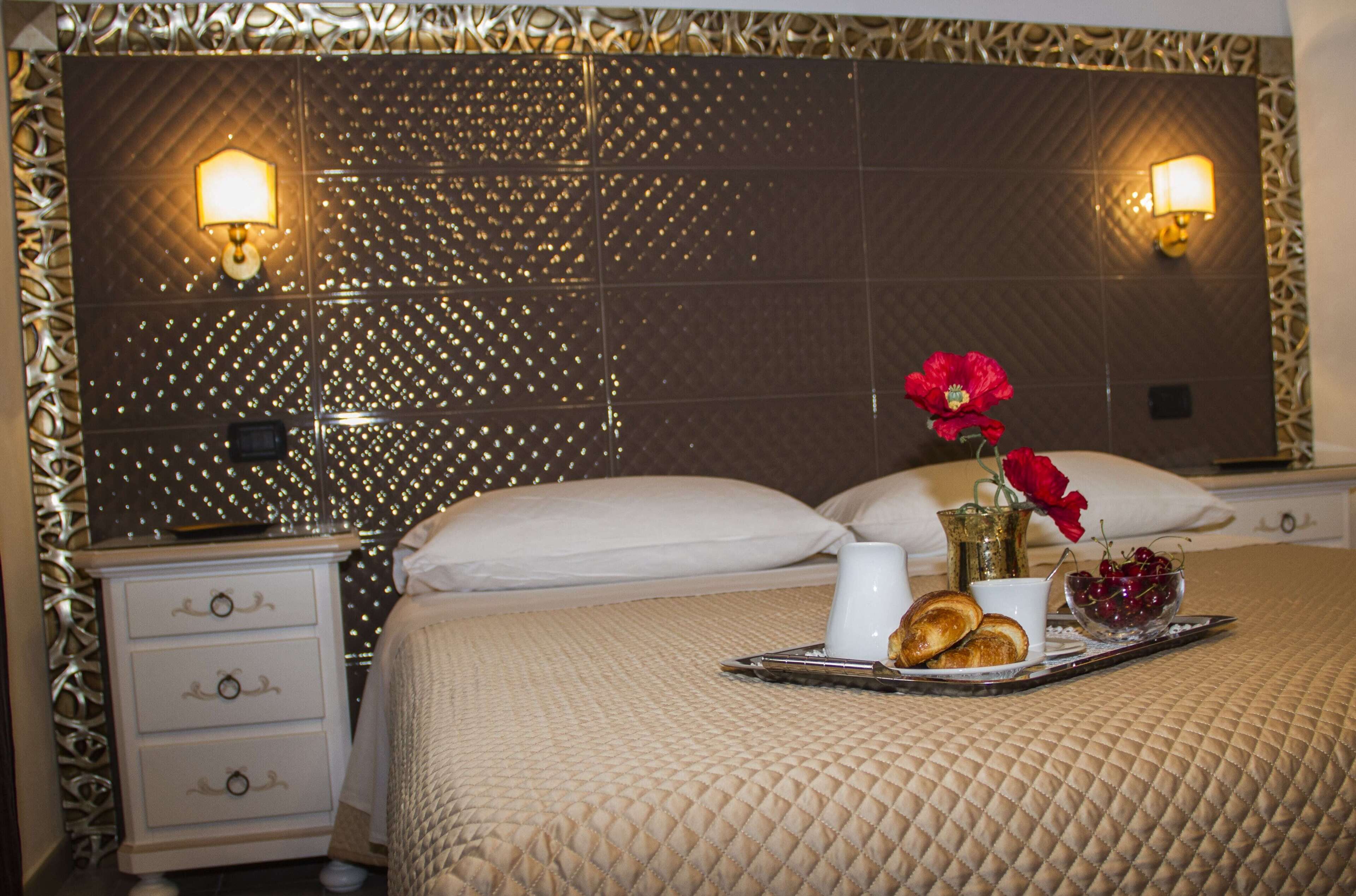 Una habitación de hotel con una cama grande, una mesita de noche, una bandeja con alimentos para el desayuno y apliques decorativos en la pared.
