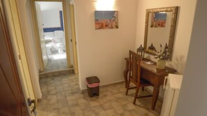 Een gang met een betegelde vloer, een houten bureau met stoel, een kleine prullenbak en een spiegel aan de muur.