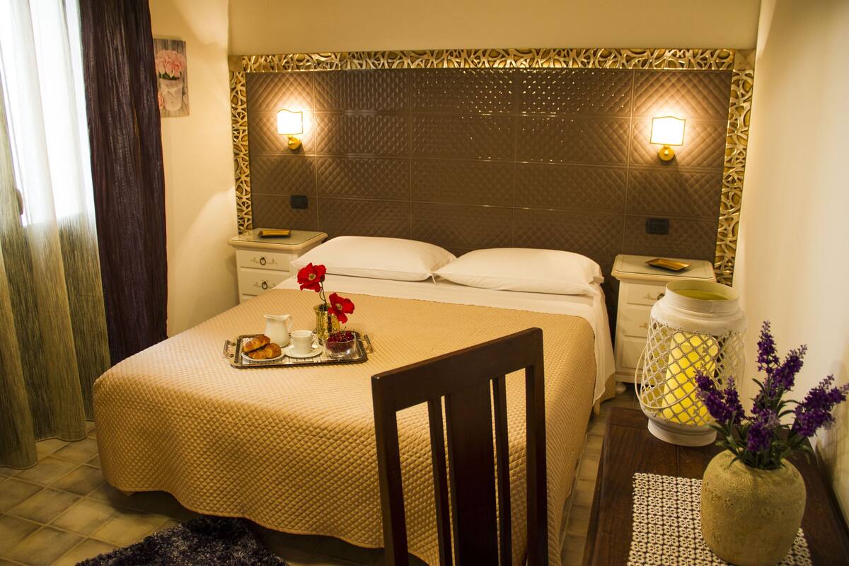 Una habitació d'hotel amb un llit, taules de nit, una cadira, un vas amb flors i un llum de llanternatge.