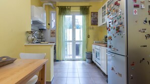 A kitchen with a wooden table, a refrigerator with magnets, and a window with green curtains. - JOIVY Cozy Flat for 4 w/ Patio, Nearby Navigli (Milan)