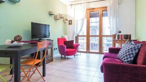 A living room with a TV, a dining table, two chairs, a sofa, and a balcony with curtains. - JOIVY Cozy Flat for 4 w/ Patio, Nearby Navigli (Milan)