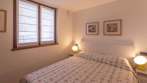 A bedroom with a bed, two bedside lamps, a window, and two framed pictures on the wall.