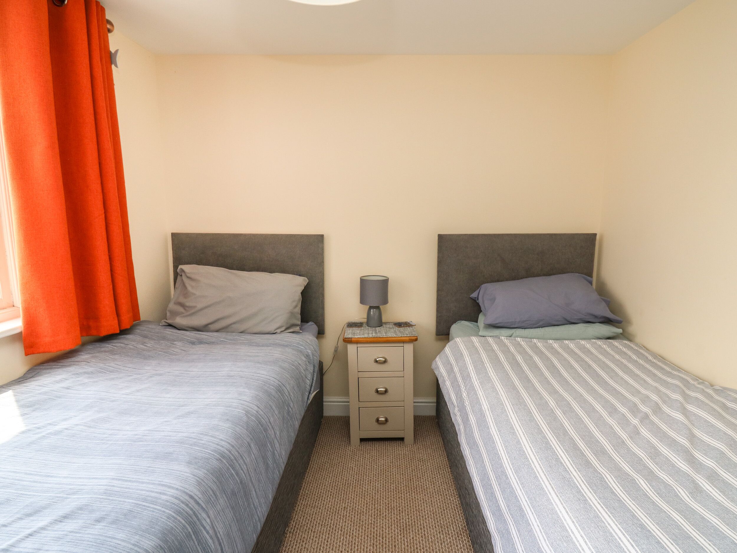 A twin room with two beds, a nightstand with a lamp, and a window with orange curtains.
