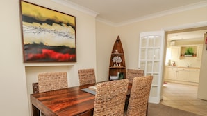 Un comedor con mesa de madera y sillas tejidas, un cuadro en la pared y una cocina visible a través de una puerta abierta.