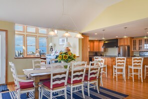 A dining area with a table and chairs, a kitchen in the background, and a view through the window. - Home On The Range: Private Pool, Hot Tub, Elevator, Village Beach Club Access (Nags Head)