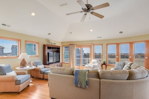 A spacious living room with a ceiling fan, large windows, and a view of the outdoors. - Home On The Range: Private Pool, Hot Tub, Elevator, Village Beach Club Access (Nags Head)
