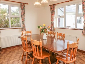 A dining room with a wooden table and chairs, a vase with flowers, and large windows with curtains. - Cleary Cottage (Miltown Malbay)