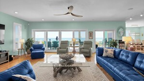 A spacious living room with a large glass coffee table, blue leather sofas, and a ceiling fan. -  Siren's Song--Gorgeous Oceanfront House with Pool and Spa! (Kure Beach)