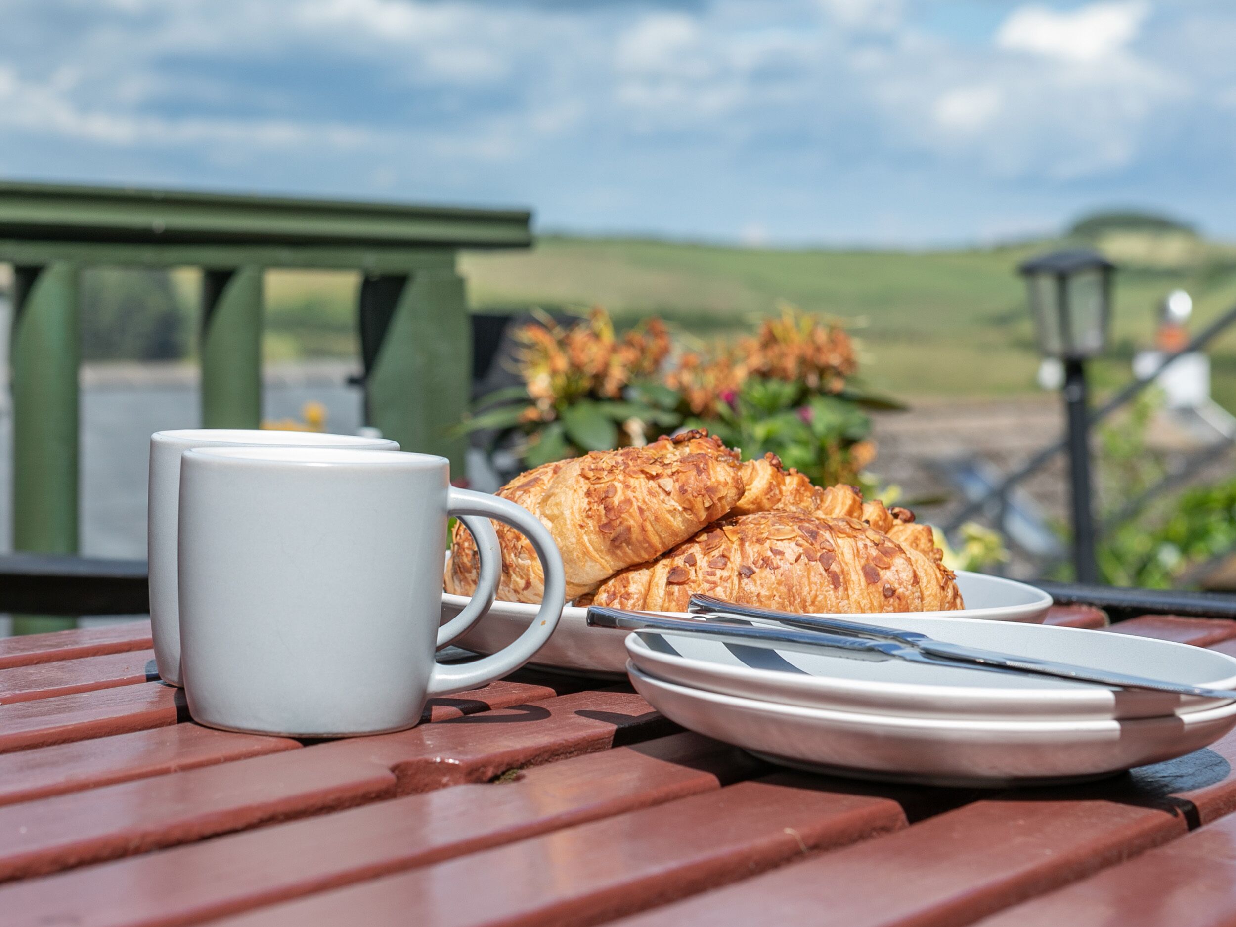 A picnic table with a white mug, a plate with a knife, and two croissants.