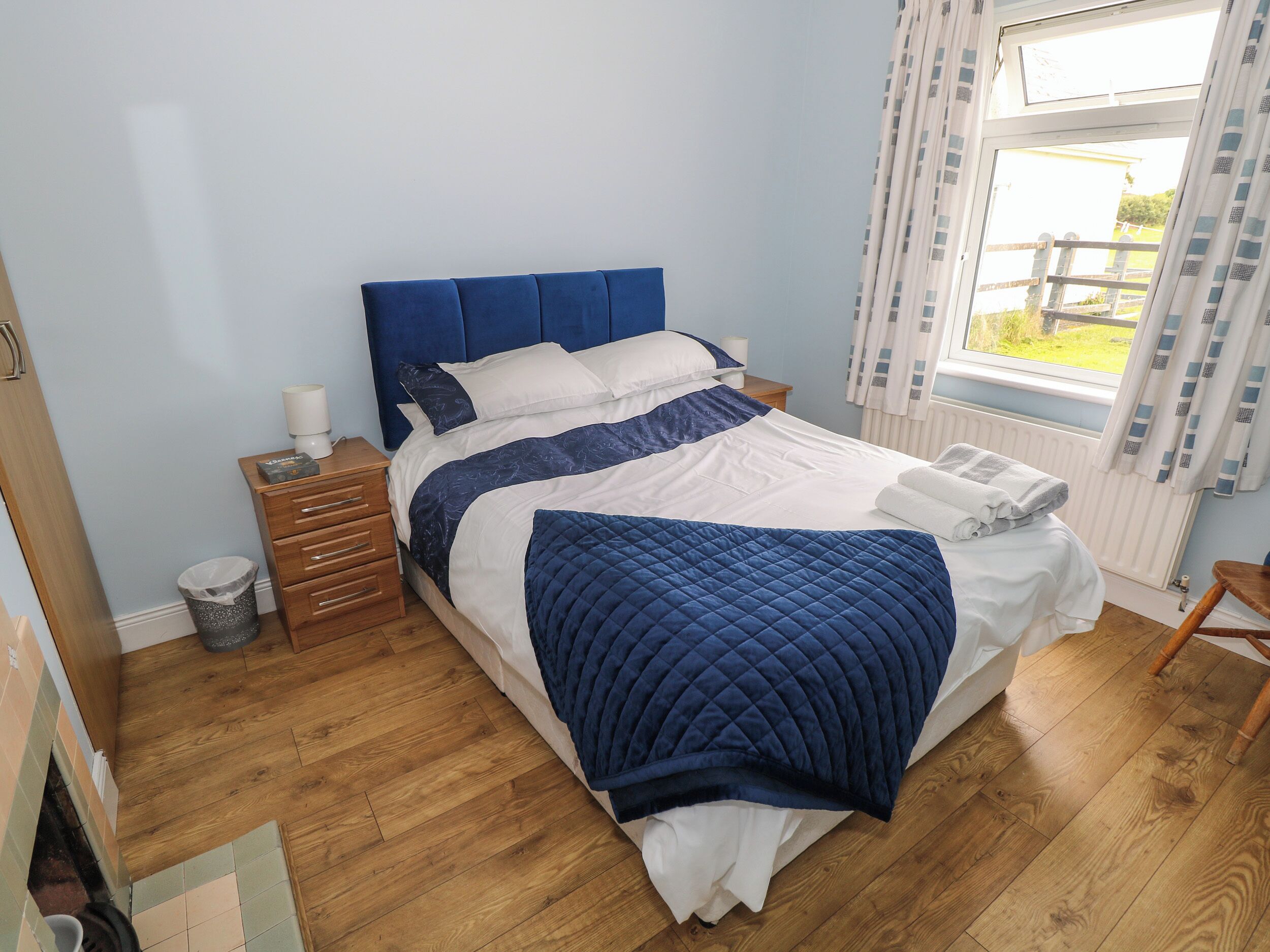 A neatly made bed with a blue headboard, a wooden nightstand with a lamp, and a view of the outdoors through a window with curtains.