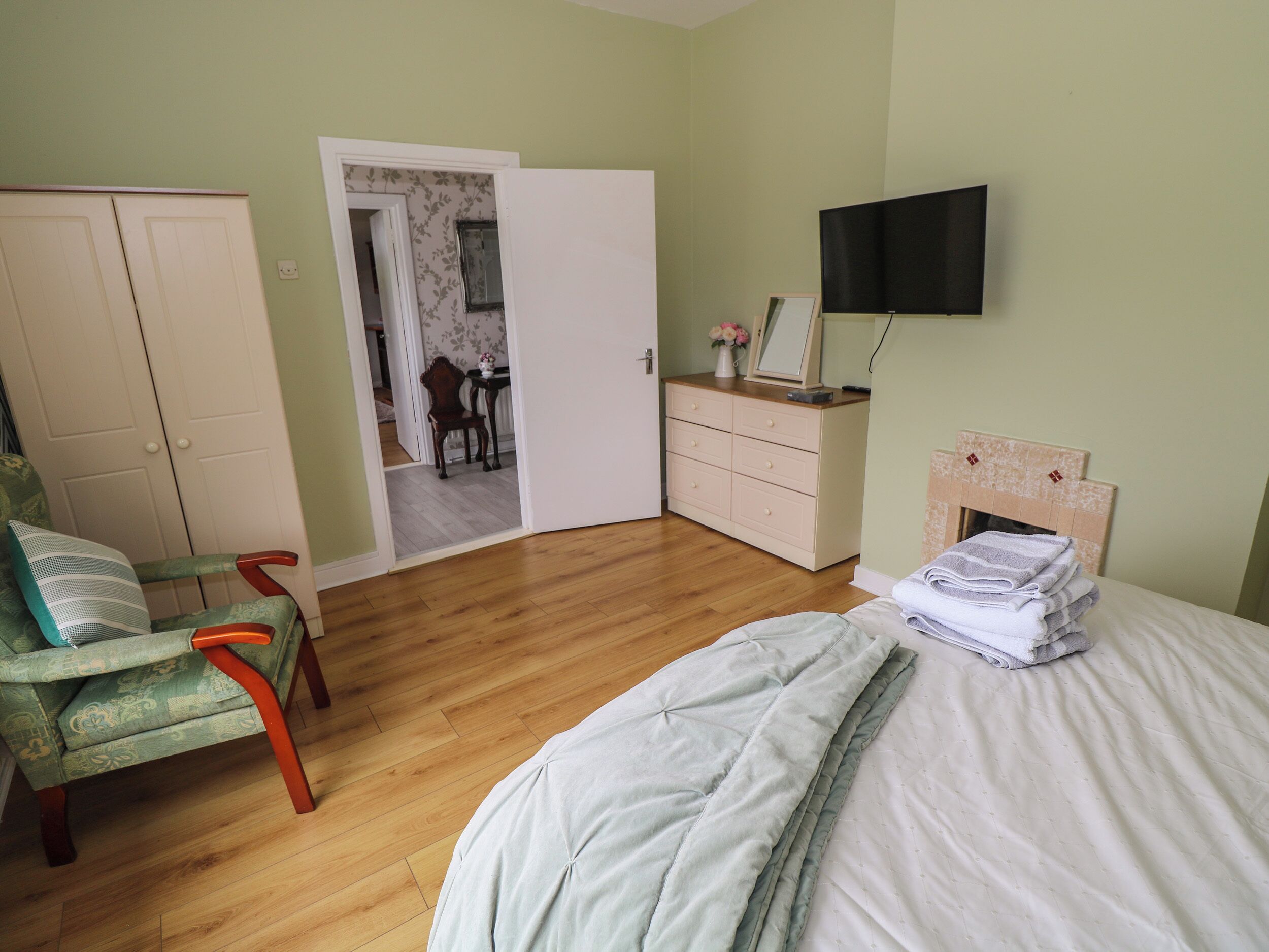 Quarto com cama, cadeira, cômoda, televisão e porta que dá para outro espaço.