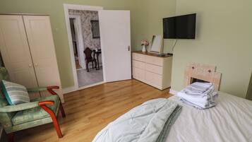 Quarto com cama, cadeira, cômoda, televisão e porta que dá para outro espaço.